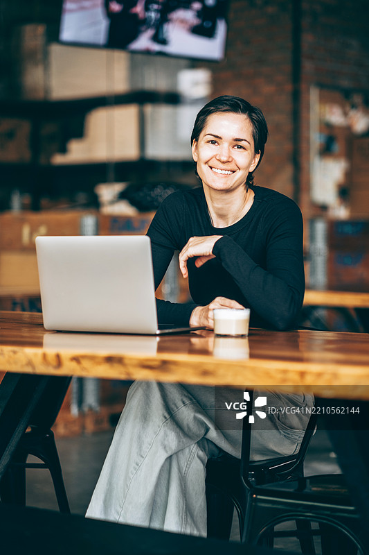 一位留着深色短发的年轻女子坐在长木桌旁，桌上放着笔记本电脑和咖啡，她正对着镜头微笑。背景中可以看到箱子和工具。图片素材