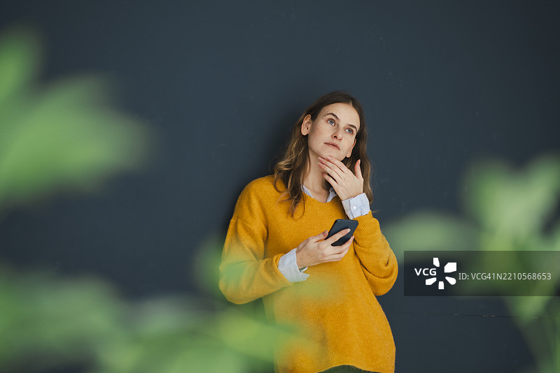 穿着黄色毛衣的沉思女性在室内拿着智能手机图片素材