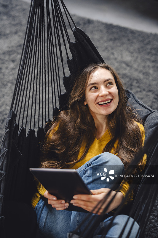 年轻女性在家中吊床上放松，手里拿着平板电脑图片素材