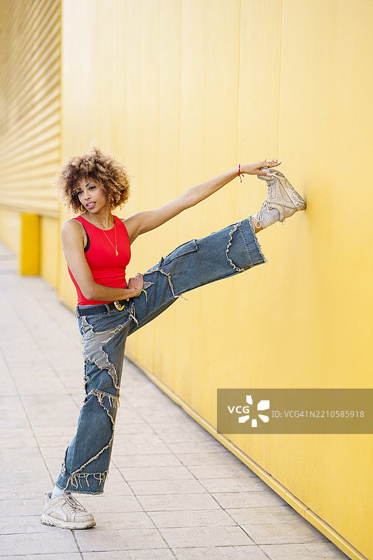 在户外的黄色墙壁前，留着卷曲非洲发型的女性正在伸展身体图片素材