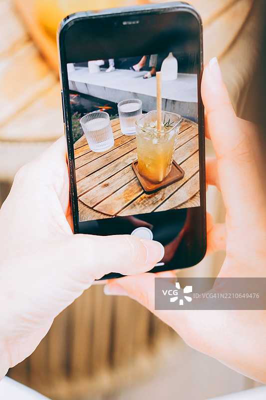 特写镜头，手握品牌智能手机拍摄木桌上的冰镇草本饮料。屏幕上显示着旁边的水杯。户外咖啡馆场景，轻松的氛围。图片素材