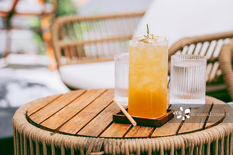 带花卉装饰的黄色冰饮料放在方形木垫上，置于圆形藤编桌上。旁边放着两个有纹路的水杯。背景柔和，有露台家具。图片素材