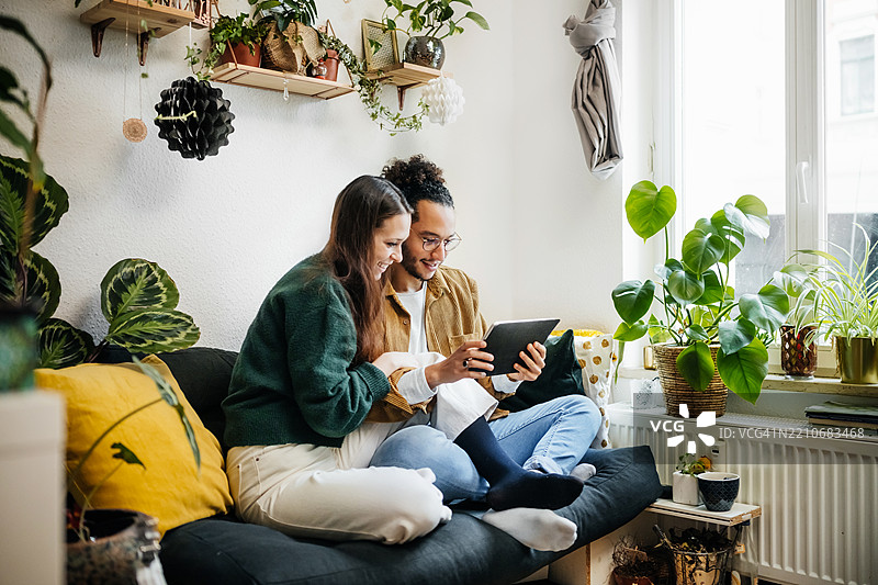 微笑的情侣坐在沙发上，周围环绕着植物，浏览平板电脑。图片素材