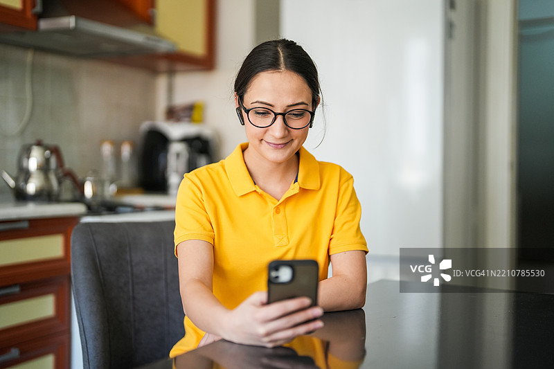 年轻女子穿着黄色T恤，坐在厨房桌旁，通过手机远程工作。图片素材