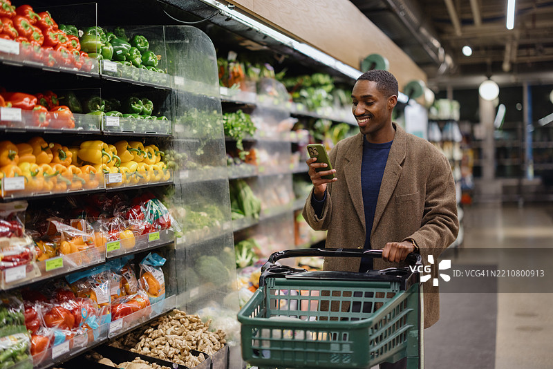 男子在超市使用智能手机购物，推着购物车靠近新鲜蔬菜。图片素材