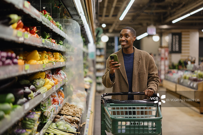 男子在杂货店购物，微笑着拿着手机挑选新鲜农产品图片素材