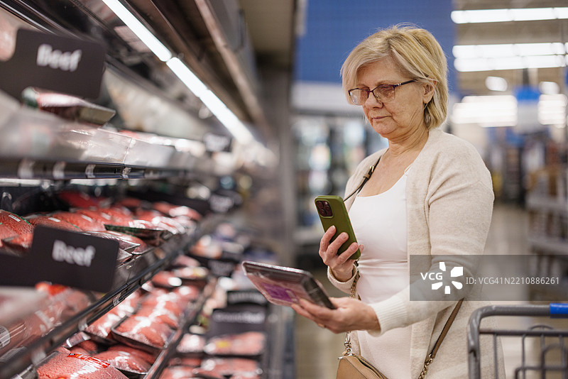 老年女性在杂货店购物时使用智能手机查看牛肉图片素材