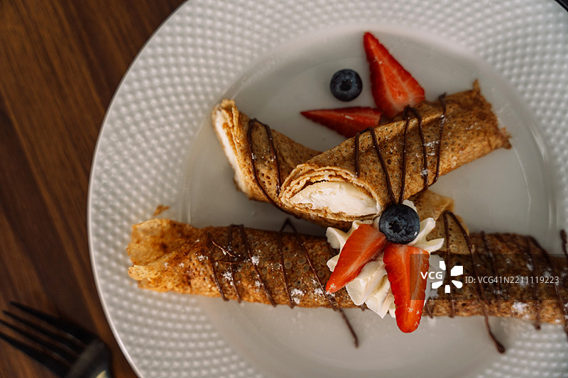 白盘上的薄煎饼卷，配有浆果、巧克力糖浆和餐具图片素材