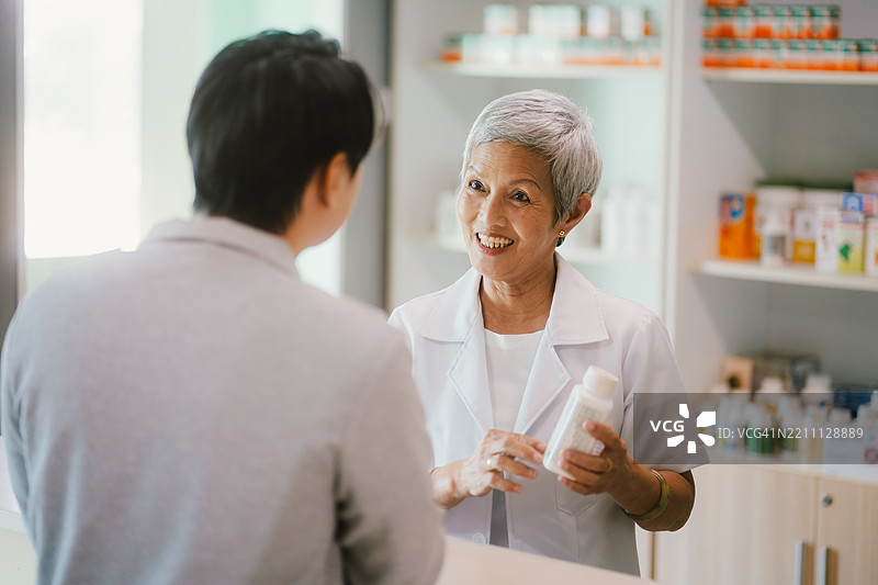 亚洲老年药剂师在药店环境中将药物递给一位男性顾客，背景是药品货架。图片素材