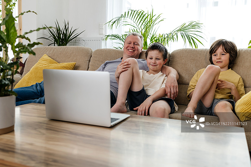 父亲和孩子们坐在家里的沙发上看笔记本电脑上的卡通，家庭一起度过美好时光。图片素材