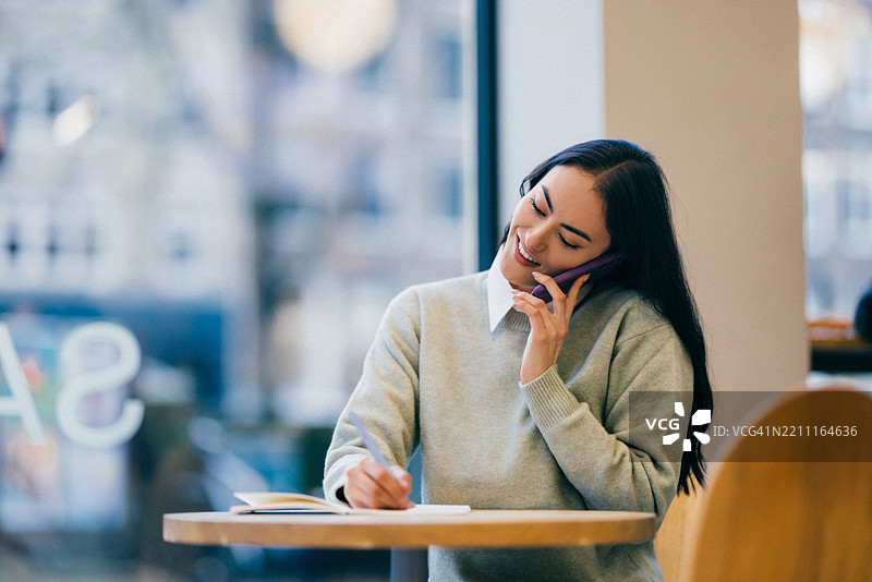 在舒适的咖啡馆里，一位职业女性正在打电话并写笔记图片素材