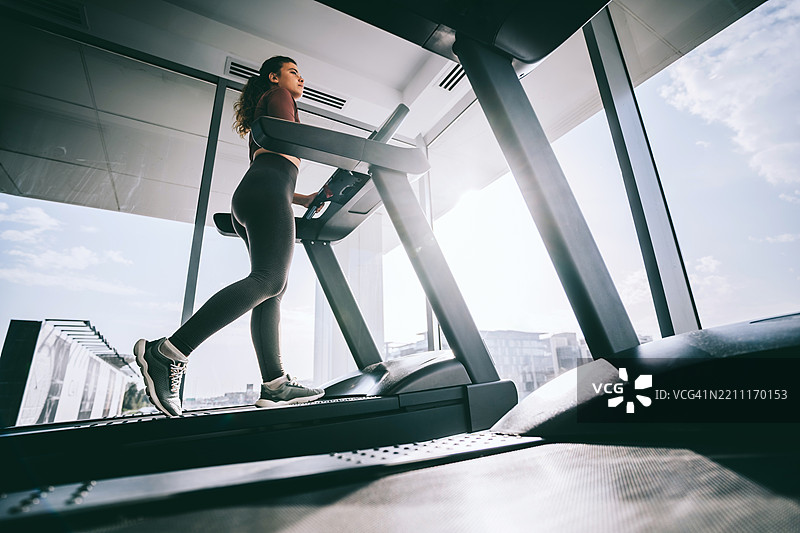 在现代健身房的大窗户旁，女性在跑步机上锻炼。图片素材