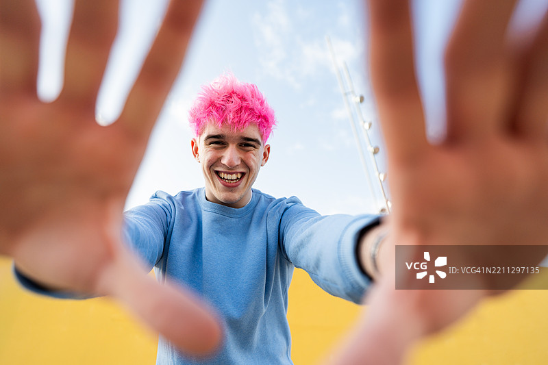 年轻男子微笑着伸出手与镜头互动，粉色头发。图片素材