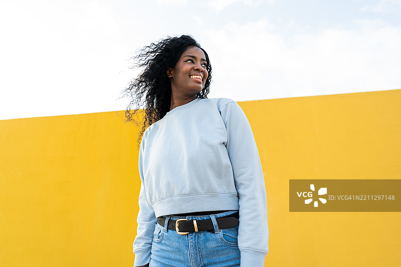 年轻的黑人女性微笑着，侧身望向黄色墙壁。图片素材