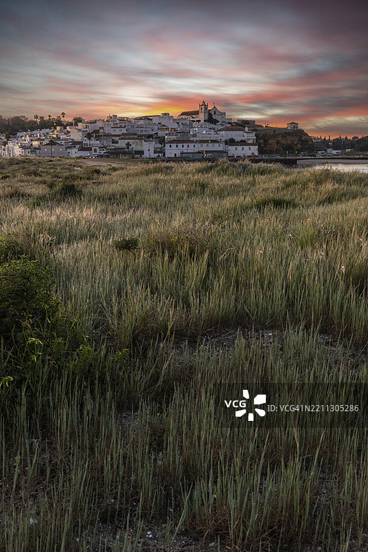 日出时分，俯瞰草地、沙滩和退潮时的港口。葡萄牙阿尔加维波尔蒂芒费拉古度的历史古镇全景图。图片素材
