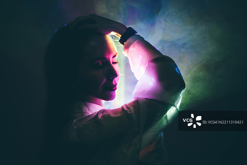 在黑暗的背景下，一位女士的侧脸被一道鲜艳的霓虹灯光和周围的彩色烟雾戏剧性地照亮，她的手触摸着头部。图片素材