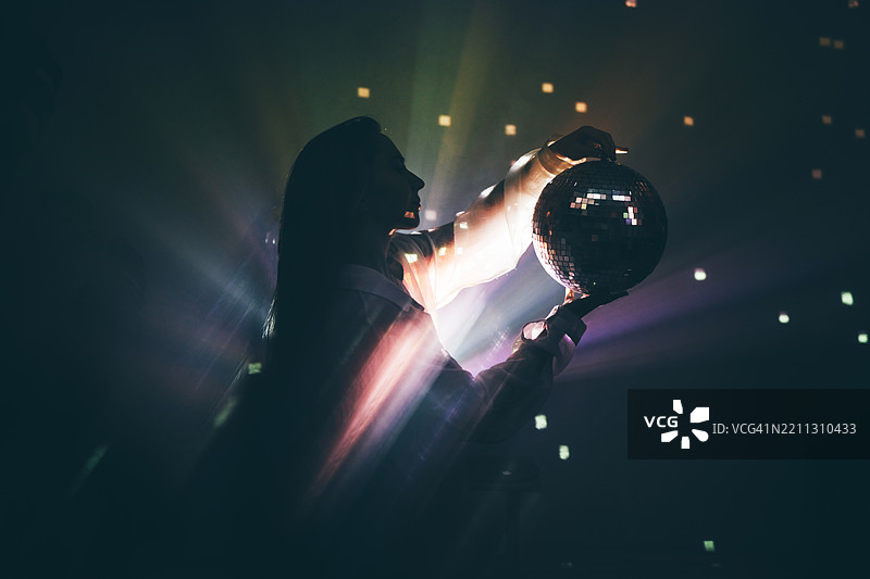 一位女士在黑暗、富有氛围的环境中手持迪斯科球，球体将光线反射到她的手和脸上。神秘、魔法与庆典。图片素材