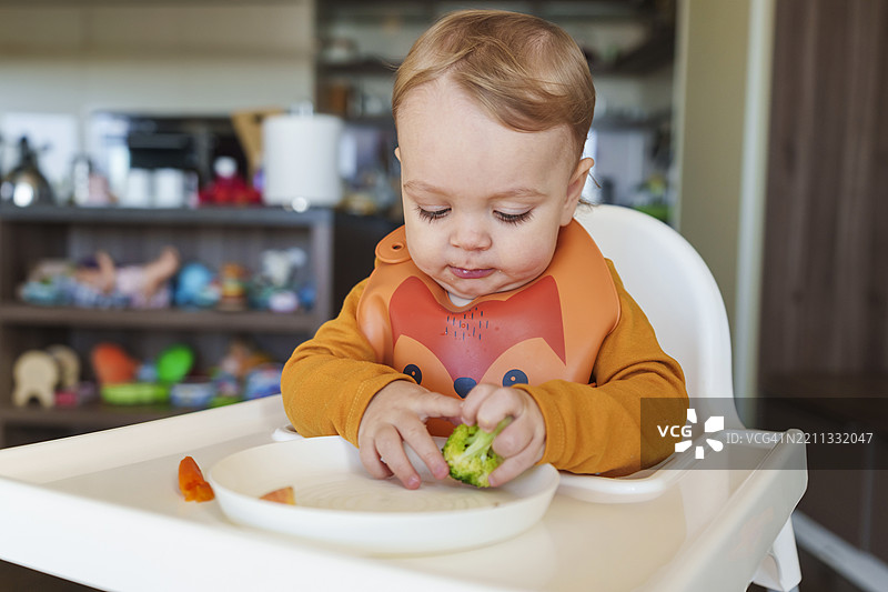可爱的小男孩坐在椅子上，手里拿着盘子里的西兰花。图片素材