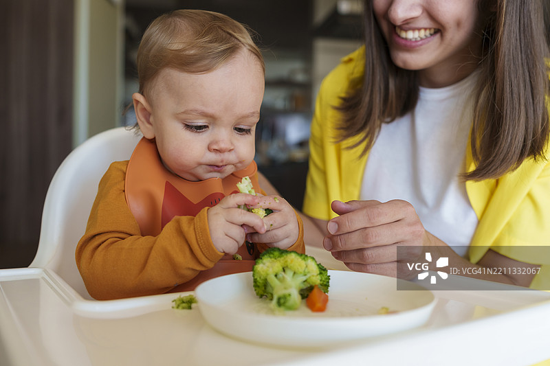 快乐的母亲在家中教儿子吃西兰花，儿子坐在高脚椅上。图片素材