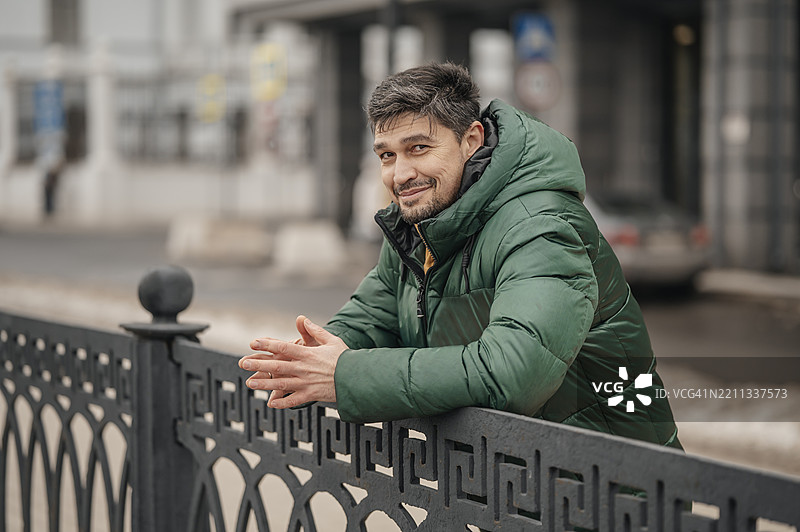 穿着绿色夹克的男子在城市中微笑着倚靠在栅栏上图片素材