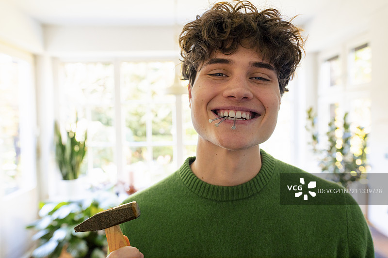 年轻男子微笑着，嘴里叼着钉子，手里拿着锤子，站在家中。图片素材