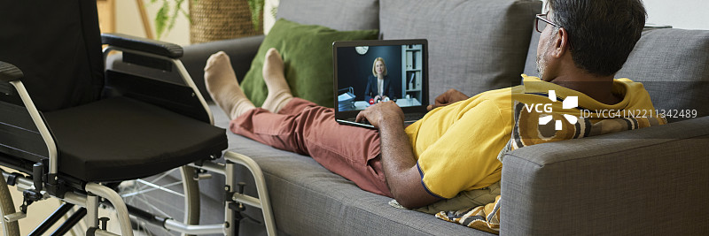 男子躺在沙发上，通过笔记本电脑进行视频通话，旁边有一把轮椅。图片素材