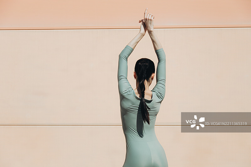 一位穿着灰色紧身衣的年轻女性背影在桃色墙壁旁摆姿势。图片素材