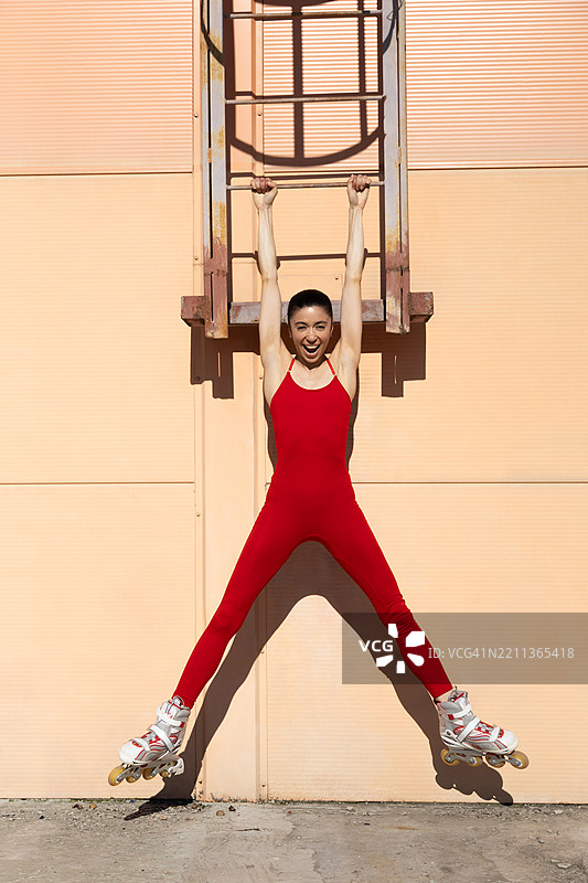 穿着红色紧身衣和滑轮鞋的快乐年轻女性悬挂在桃色墙壁上的梯子上。图片素材