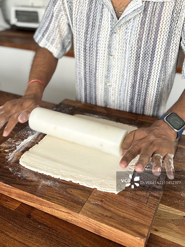 在家厨房中，一位无法辨认的男子正在使用大理石擀面杖将一块酥皮面团擀平，木砧板上，画面为俯视角度，重点在前景，家庭烹饪概念。图片素材