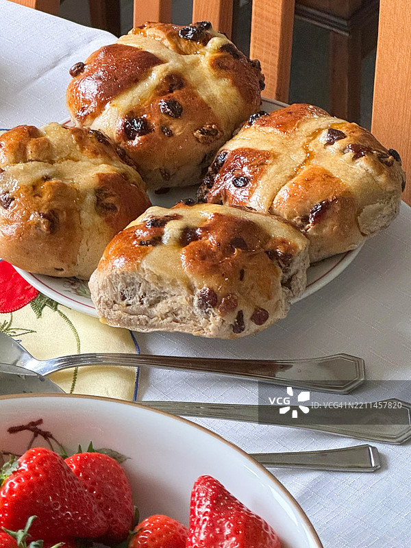 复活节自助餐的家庭餐桌特写，盘子里放着热十字面包，碗里装着草莓，不锈钢餐具，白色桌布，家庭环境，俯视角度，重点在前景。图片素材