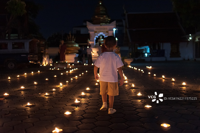 一个无法辨认的孩子在泰国的水灯节夜晚参观寺庙，地上点燃着蜡烛。图片素材