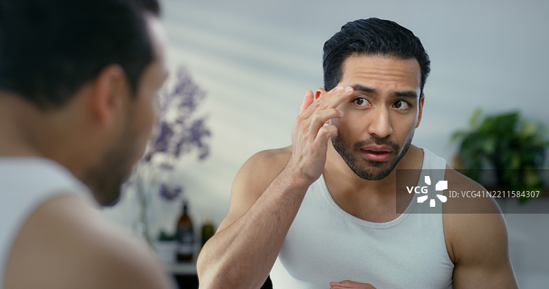 亚洲男性在家浴室中使用护肤品照镜子，进行美容、修饰或早晨护理。男性正在检查眉毛，注重面部或皮肤护理的卫生。图片素材