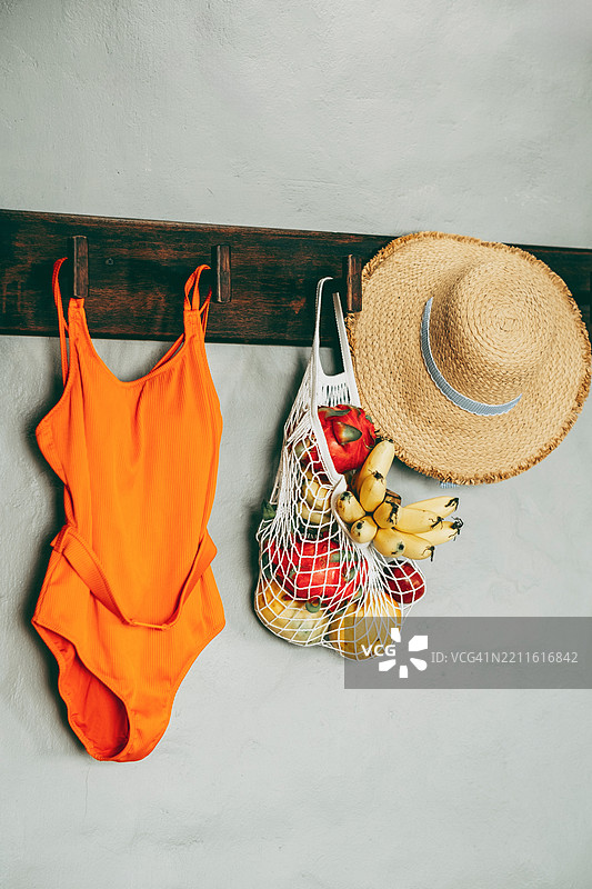 橙色泳衣、草帽和装满新鲜水果的网袋挂在浅色墙壁上的木钩上。图片素材