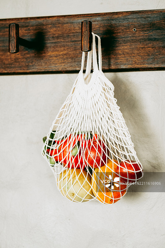 白色网袋里装满了芒果、火龙果和百香果，挂在浅色墙壁上的木钩上。图片素材