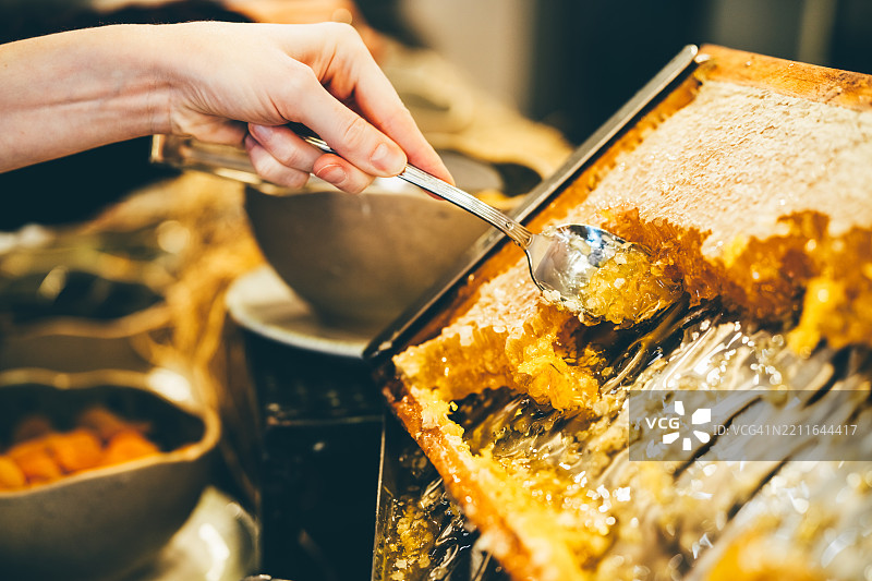 一只手在自助餐上，用一把银勺从大的木制蜂巢架中舀取新鲜的金黄色蜂蜜。这张特写照片突出了蜂蜜的有机质地以及从农场到餐桌的天然奢华概念。图片素材