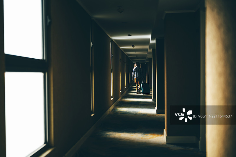 一名旅客拉着行李箱，走向走廊尽头的自然光，走在一条昏暗的长酒店走廊里。图片素材