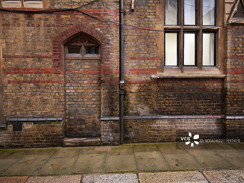 风化且肮脏的棕色砖墙，封闭的门和亮着的窗户。电缆。铺砌的人行道。伦敦，英格兰，联合王国。图片素材