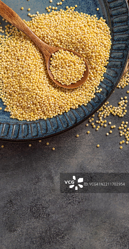 蓝色盘子里装着生的黄色去壳小米，旁边有一把木勺，灰色桌面俯视图。图片素材
