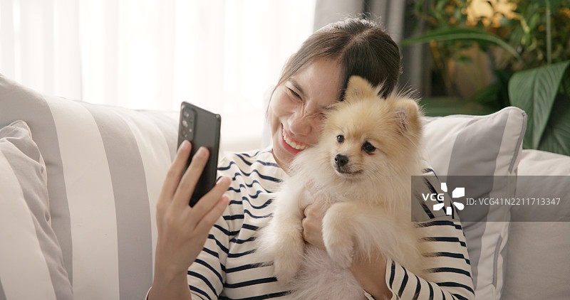 亚洲女性在家里的沙发上微笑着与可爱的小狗自拍。图片素材