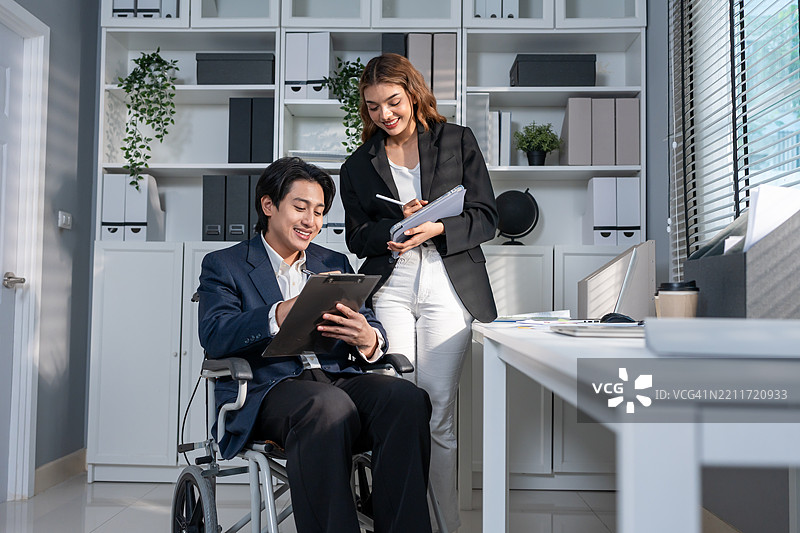 亚洲商人在办公室里使用轮椅与同事合作。年轻的员工伙伴感到快乐和放松，享受讨论项目和检查文件的时光。图片素材