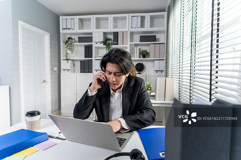 亚洲忙碌的商人一边打电话一边使用笔记本电脑在办公室工作。吸引人的男性员工坐在桌子上，感到压力和疲惫，然后在公司工作场所用手机沟通。图片素材