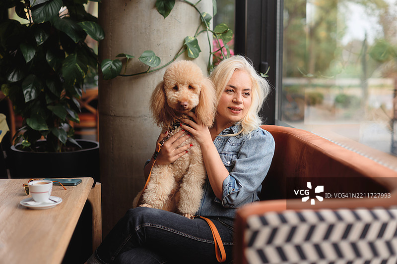 一位五十多岁的美丽女性坐在咖啡店的包厢里，怀里抱着她的棕色贵宾犬。图片素材