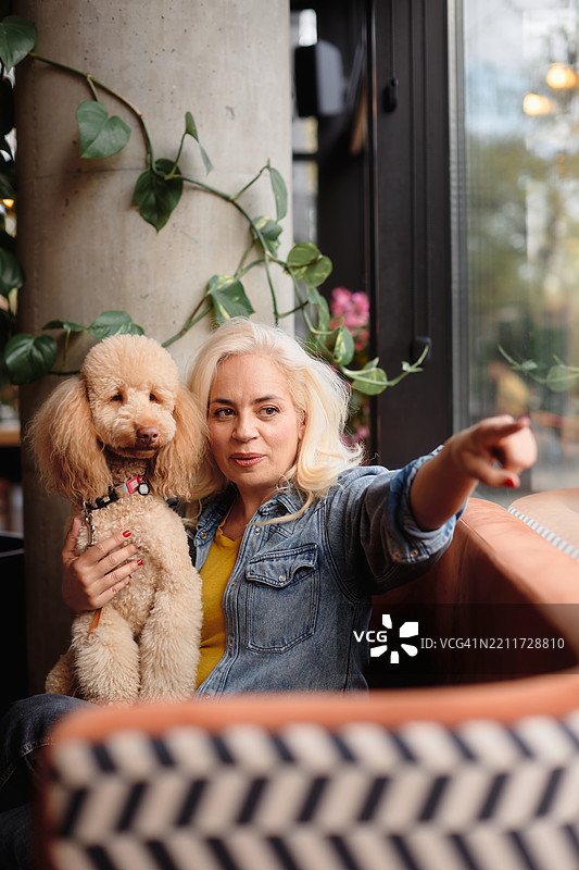 一位五十多岁的美丽女性坐在咖啡店的包厢里，怀里抱着她的棕色贵宾犬。图片素材