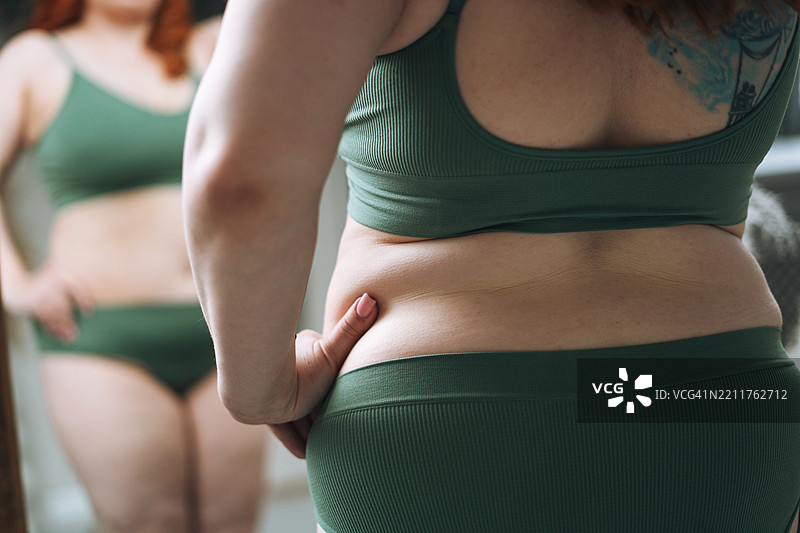 自信的红发丰满年轻女性在家卧室的镜子前穿着内衣。图片素材