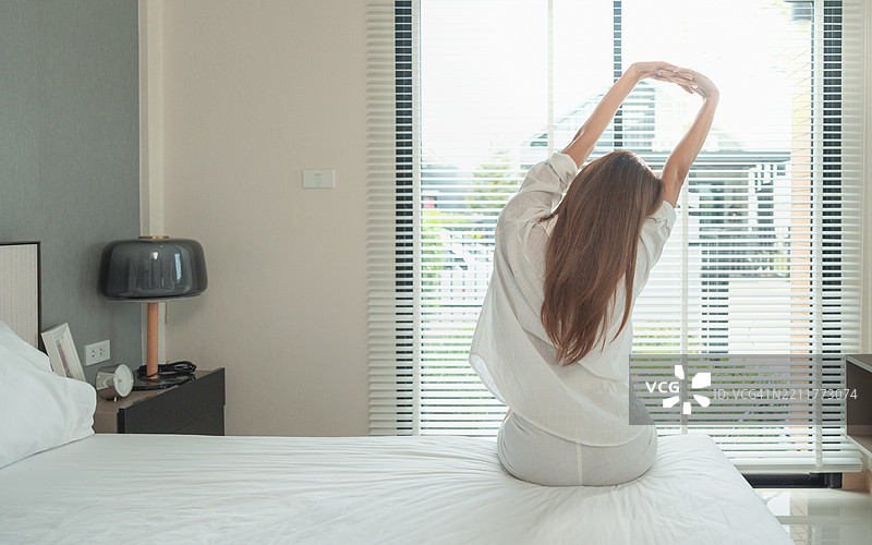 亚洲女性在家卧室起床伸展，放松身心。清晨早起，阳光明媚，感到快乐和清新。背影。图片素材