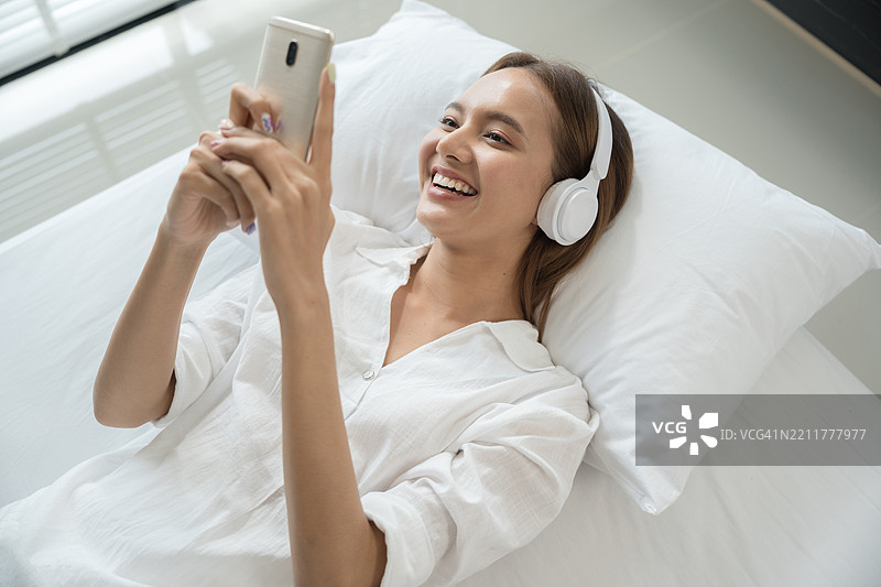 快乐的亚洲年轻女性在家中床上放松，戴着耳机观看智能手机上的视频。快乐的休闲美女在床上使用手机。图片素材