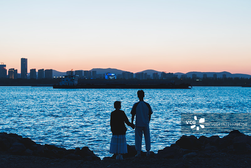 一对二十多岁的亚洲情侣在河边约会，手牵手欣赏户外的日落。图片素材