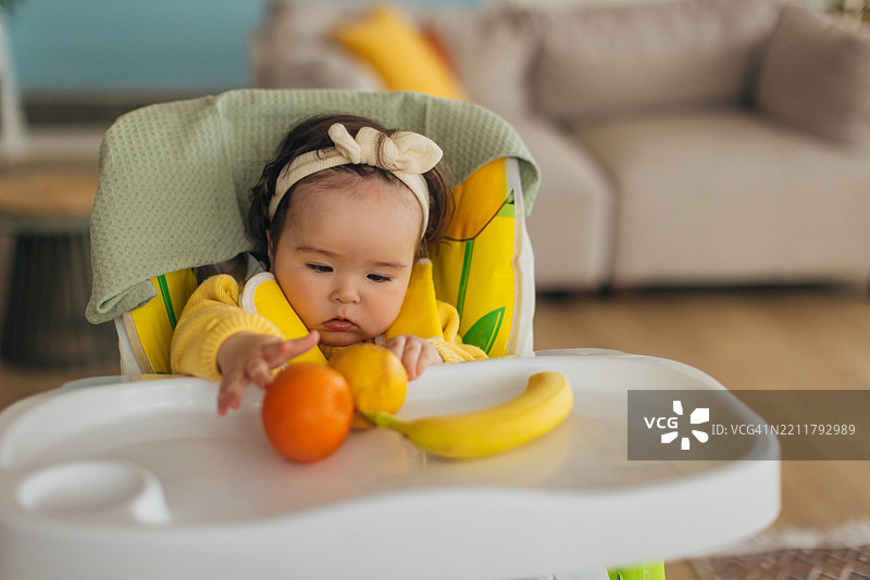 一个可爱的小多种族女孩坐在高脚椅上，开心地玩着水果。图片素材