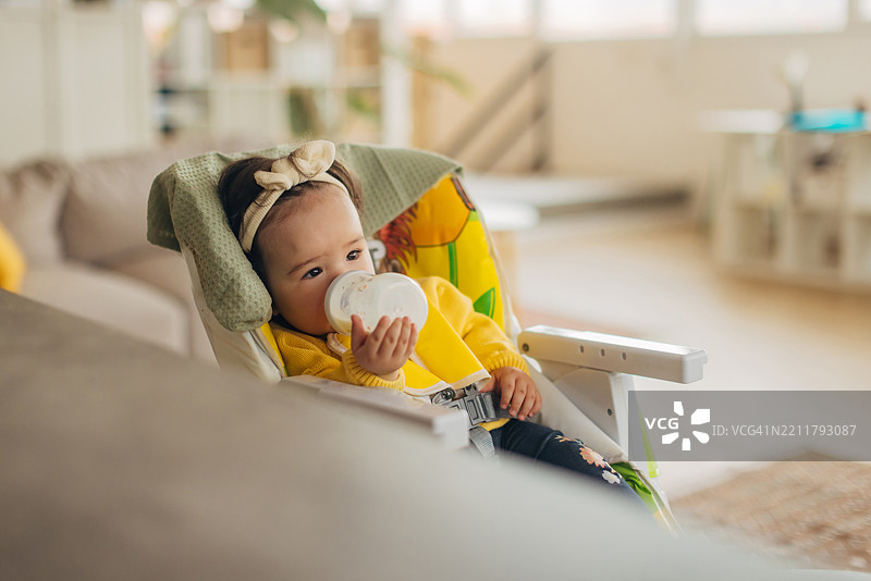 可爱的多种族婴儿女孩在高脚椅上喝着奶瓶里的牛奶，放松身心。图片素材
