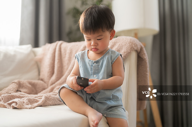 好奇的亚洲男婴拿着遥控器，展现出他与生俱来的好奇心和独立性。可爱的亚洲男孩在客厅沙发上玩电视遥控器，阳光明媚。图片素材